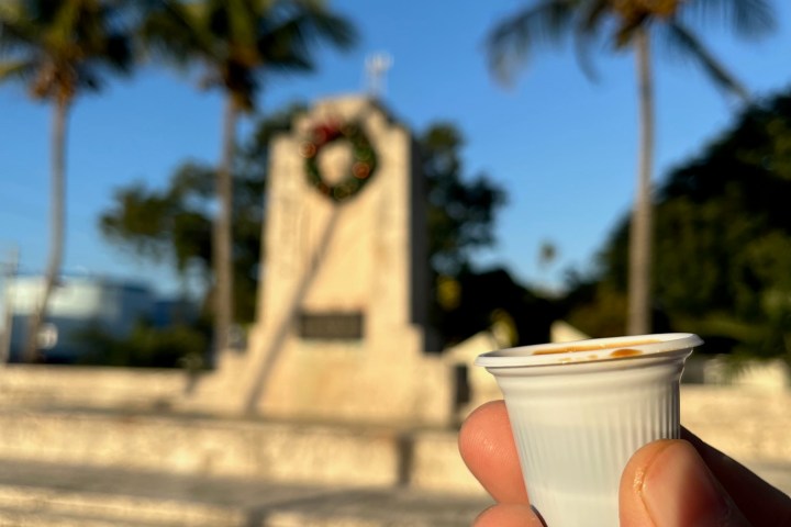 a hand holding a colada cuban coffee in front of hurricane monument in Islamorada front of a palm tree