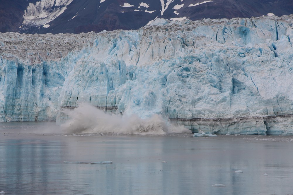 Ice calving from a glacier into the sea with mountains in the background.