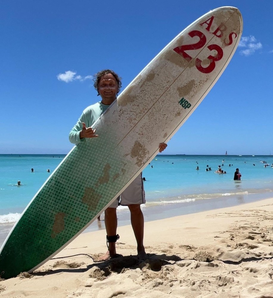 Person holding surfboard on a sandy beach with clear blue water and people swimming in the background. in Waikiki, Oahu, Hawaii