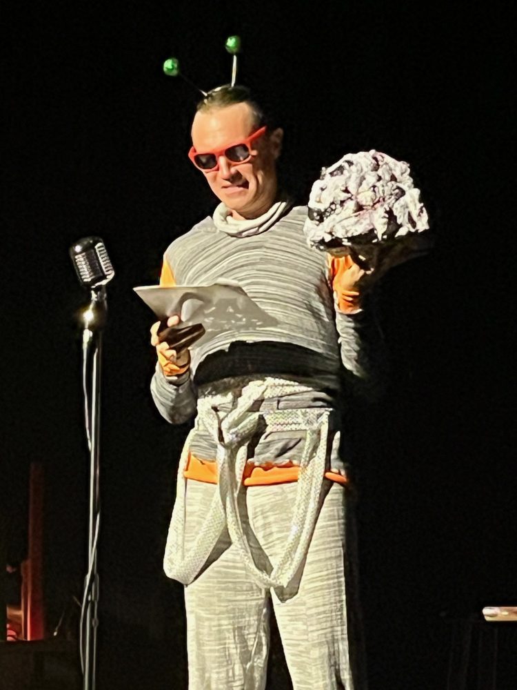 Person in costume with antennae and glasses, holding paper and prop, standing at a microphone on stage.