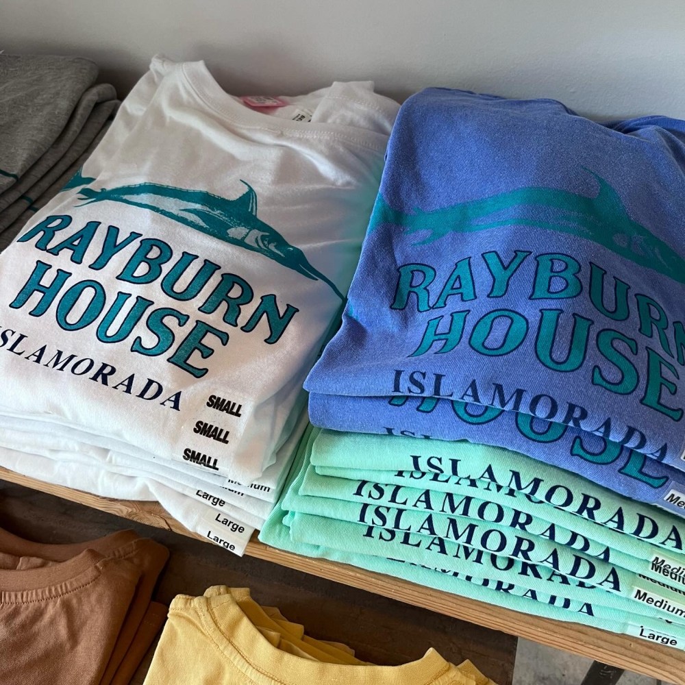 Stacked T-shirts in various colors with 'Rayburn House Islamorada' design displayed on shelves. from Netflix Bloodline Series