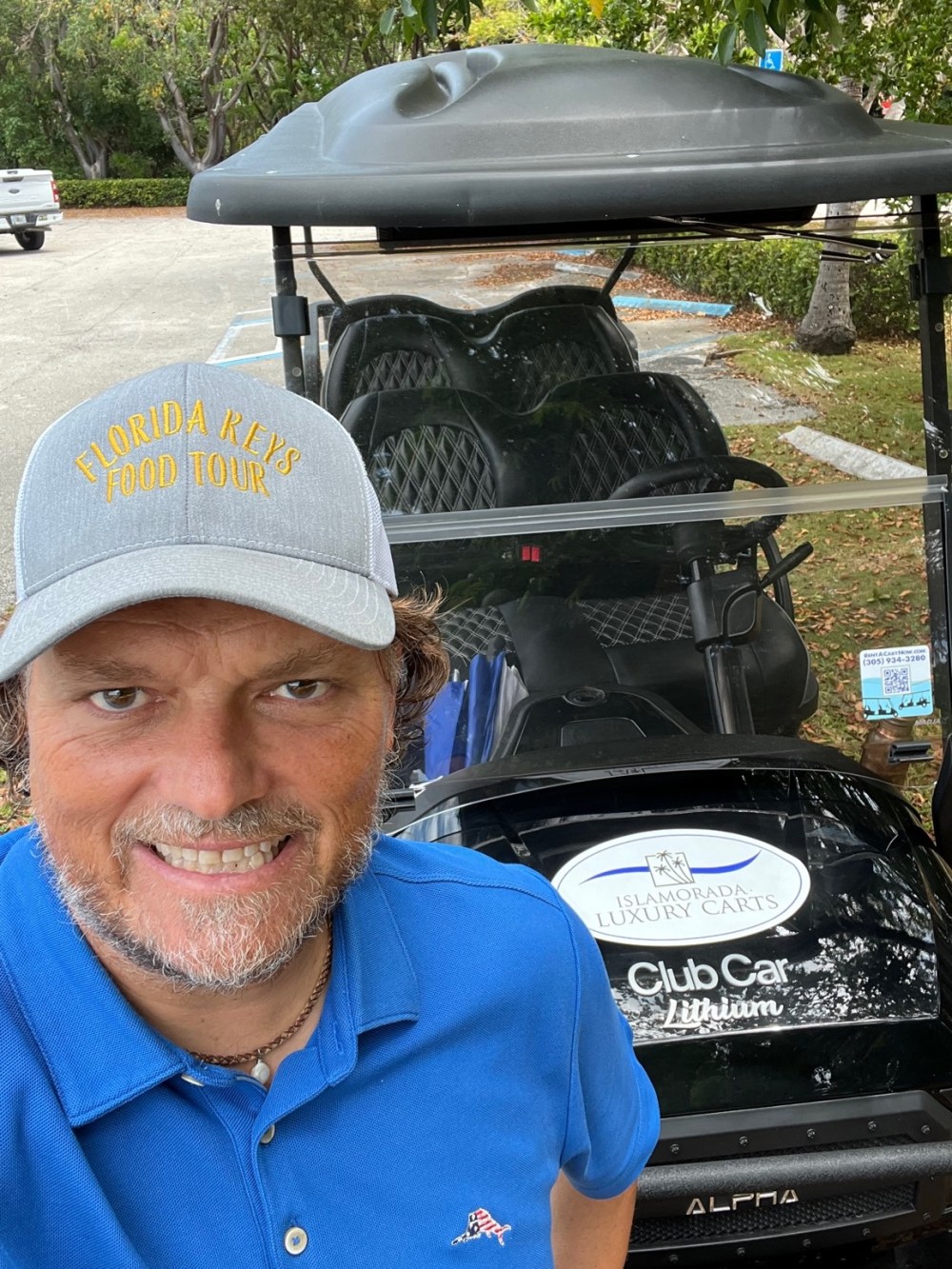 Man in blue shirt and cap smiling in front of a golf cart in a parking lot.