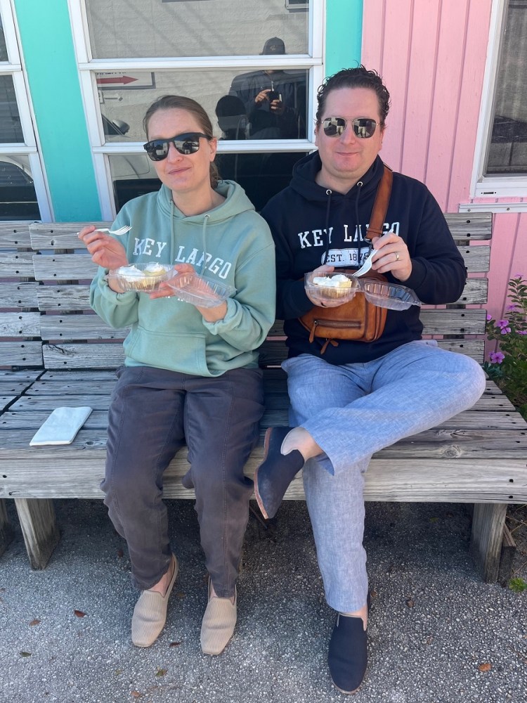 Two people sitting on a bench, eating fKey Lime Pie outside Mrs Macs wearing sunglasses and Key Largo hoodies.