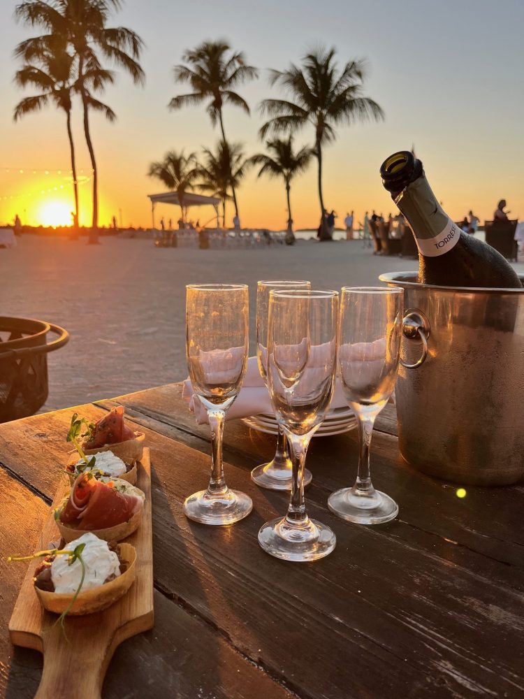 Sunset beach table with champagne, glasses, and appetizers, palm trees in background.