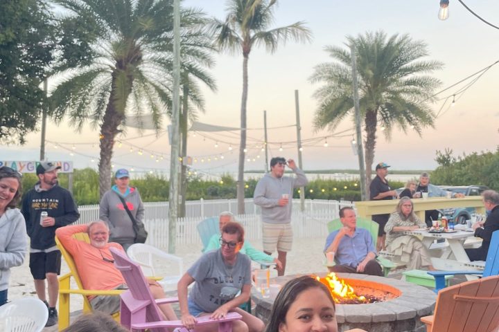 Group of people sitting around a fire pit with palm trees and string lights in the background.