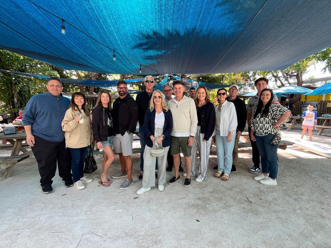 Group of people standing under a blue canopy at an outdoor venue with picnic tables.