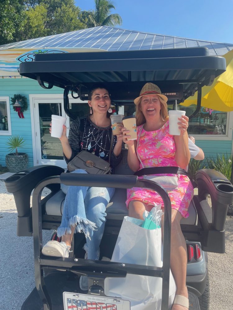 Two women sitting on a luxury golf cart holding drinks outside a boutique in islamorada smiling, sunny day.