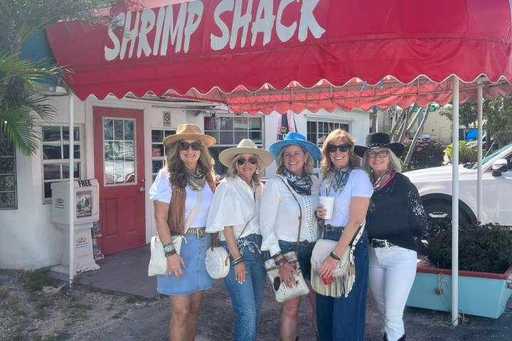 Five women in hats pose by a red awning saying 'Shrimp Shack.'