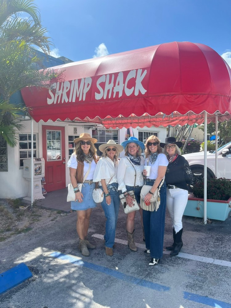 Five women in hats pose by a red awning saying 'Shrimp Shack.'